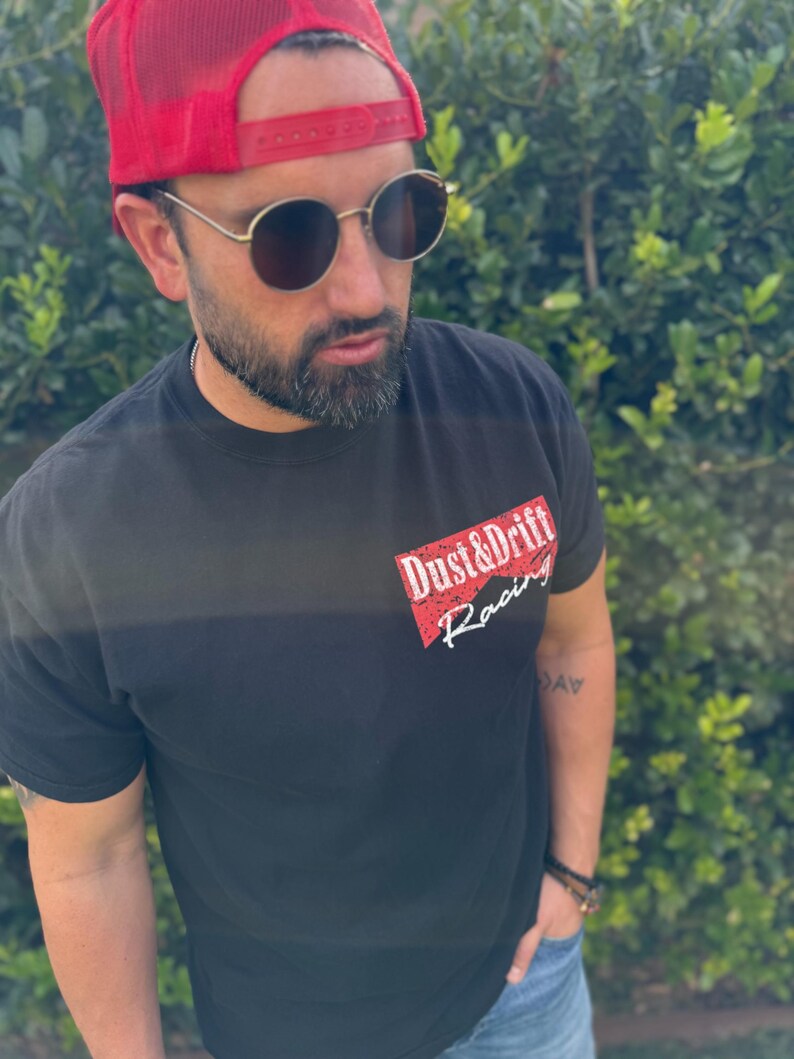 May include: Black t-shirt with the text "Dust & Drift Racing" in red and white. The person in the image is wearing a red trucker hat and round sunglasses.