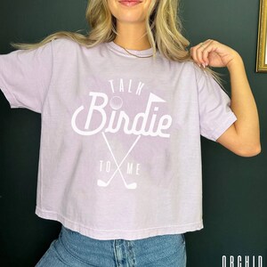 May include: A lavender-colored cropped t-shirt with a white graphic that says "Talk Birdie To Me" with a golf ball and crossed golf clubs.