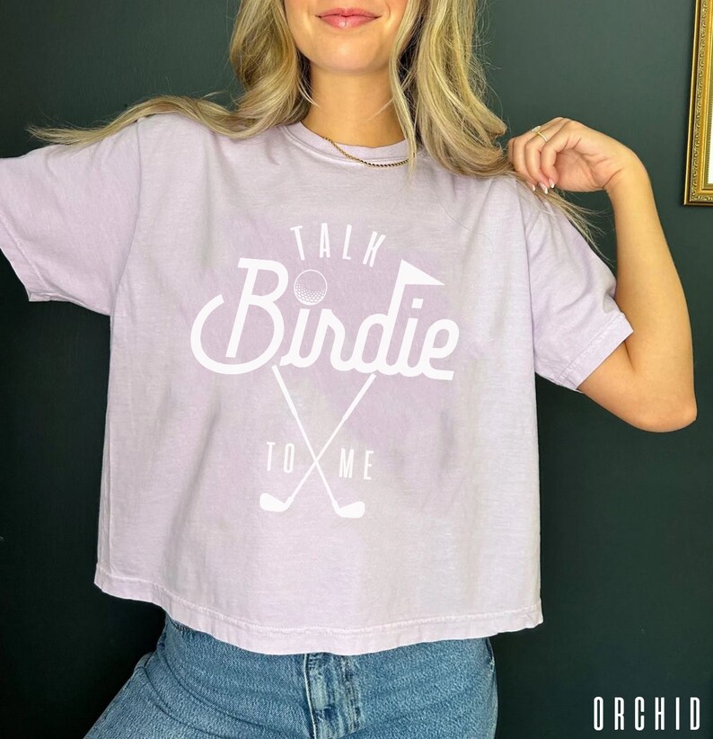 May include: A lavender-colored cropped t-shirt with a white graphic that says "Talk Birdie To Me" with a golf ball and crossed golf clubs.