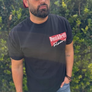 May include: Black t-shirt with the text "Dust & Drift Racing" in red and white. The shirt is short-sleeved and worn with blue jeans and a red mesh baseball cap. The person is also wearing sunglasses.