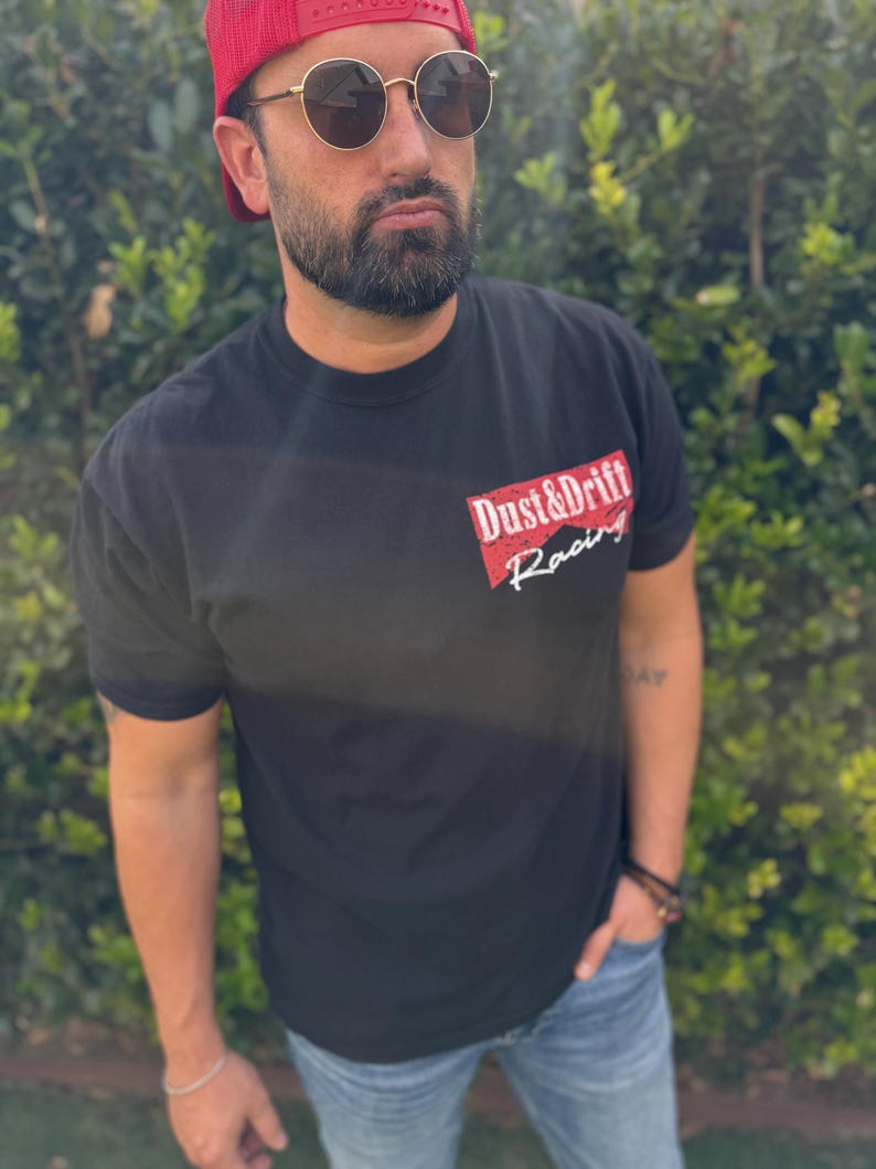 May include: Black t-shirt with the text "Dust & Drift Racing" in red and white. The shirt is short-sleeved and worn with blue jeans and a red mesh baseball cap. The person is also wearing sunglasses.