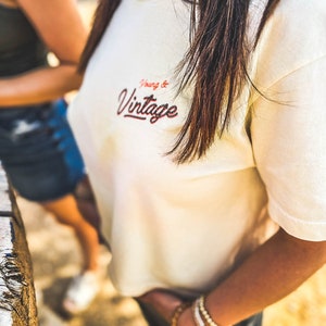 May include: Cream-colored t-shirt with the embroidered text "Young & Vintage" in red and black. The shirt has short sleeves and a crew neck. The person is wearing gold and white bracelets.