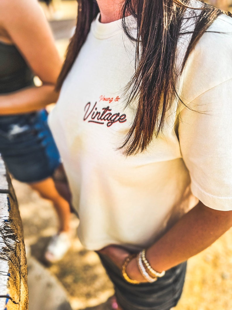 May include: Cream-colored t-shirt with the embroidered text "Young & Vintage" in red and black. The shirt has short sleeves and a crew neck. The person is wearing gold and white bracelets.