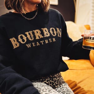 May include: A woman wearing a black sweatshirt with the text "Bourbon Weather" in gold lettering. She is holding a glass of bourbon in her right hand. She is wearing leopard print pants.