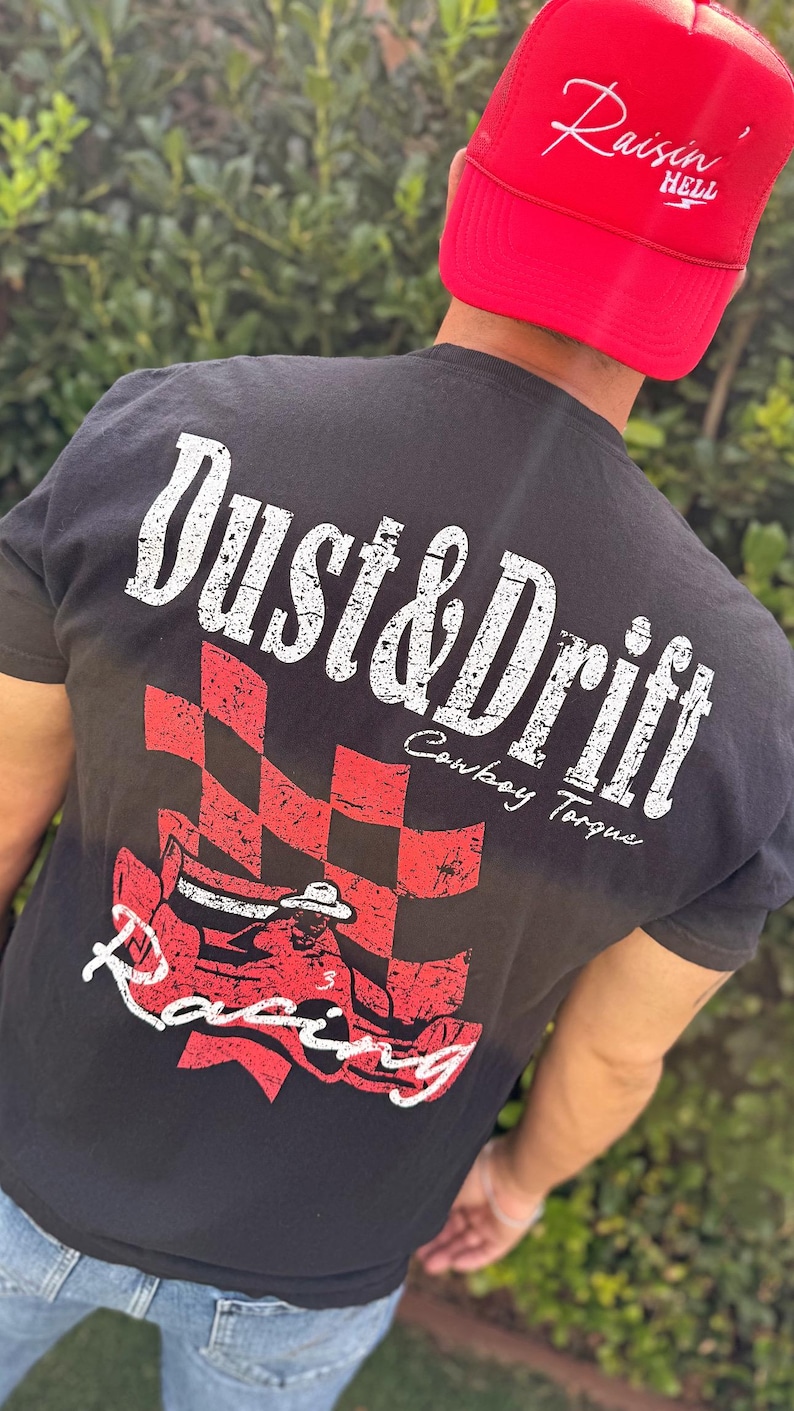 May include: Black t-shirt with a distressed white and red graphic. The graphic includes the words "Dust & Drift Cowboy Torque Racing" and a checkered flag design. A red trucker hat with the words "Raisin' Hell" is also visible.