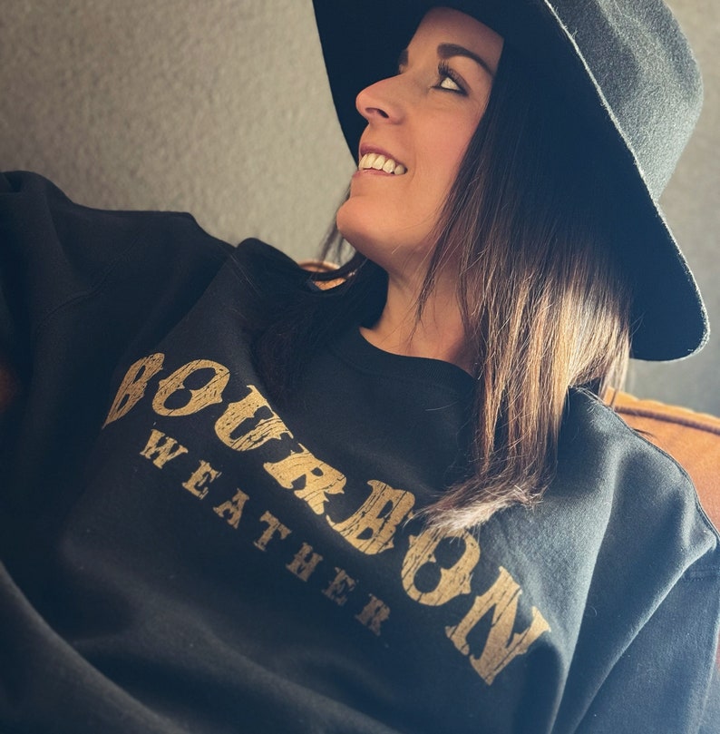 May include: A black sweatshirt with the text "Bourbon Weather" printed in gold on the front. The sweatshirt is worn by a person wearing a black hat.