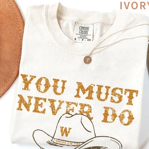 May include: Ivory t-shirt with a distressed brown graphic that says "You must never do" with a cowboy hat and the text "I guess I'm the" underneath.
