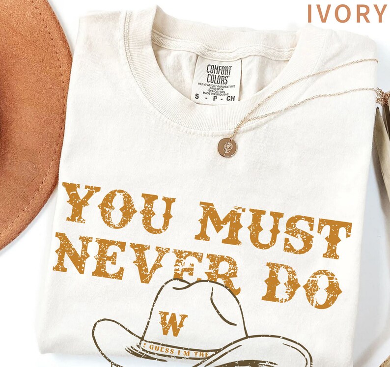 May include: Ivory t-shirt with a distressed brown graphic that says "You must never do" with a cowboy hat and the text "I guess I'm the" underneath.