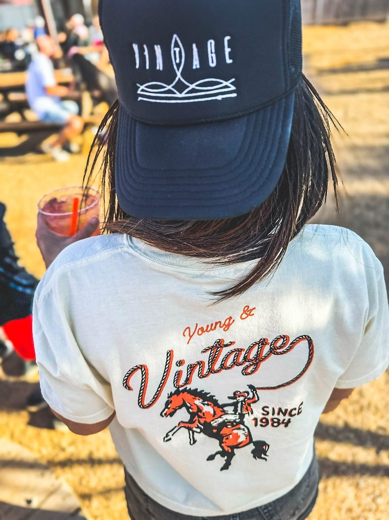 May include: A black trucker hat with the word "VINTAGE" in white, and a cream-colored t-shirt with a graphic of a cowboy on a horse and the words "Young & Vintage SINCE 1984".