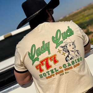 Puede incluir: Una camiseta beige con una ilustraci&oacute;n en blanco y negro de una mujer con un sombrero de vaquero. El texto "Lady Luck 777 Hotel + Casino Don't Push Your Luck Too Go." est&aacute; impreso en la parte posterior de la camiseta.