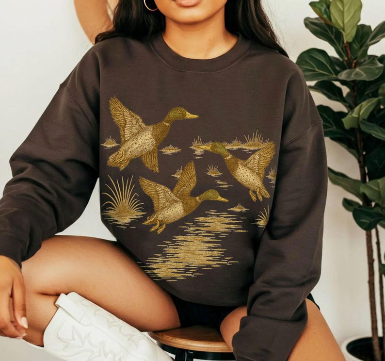 May include: A dark brown sweatshirt featuring a detailed illustration of three ducks in flight over a body of water. The ducks are depicted with brown, tan, and green coloring, with the water and reeds in a golden hue.