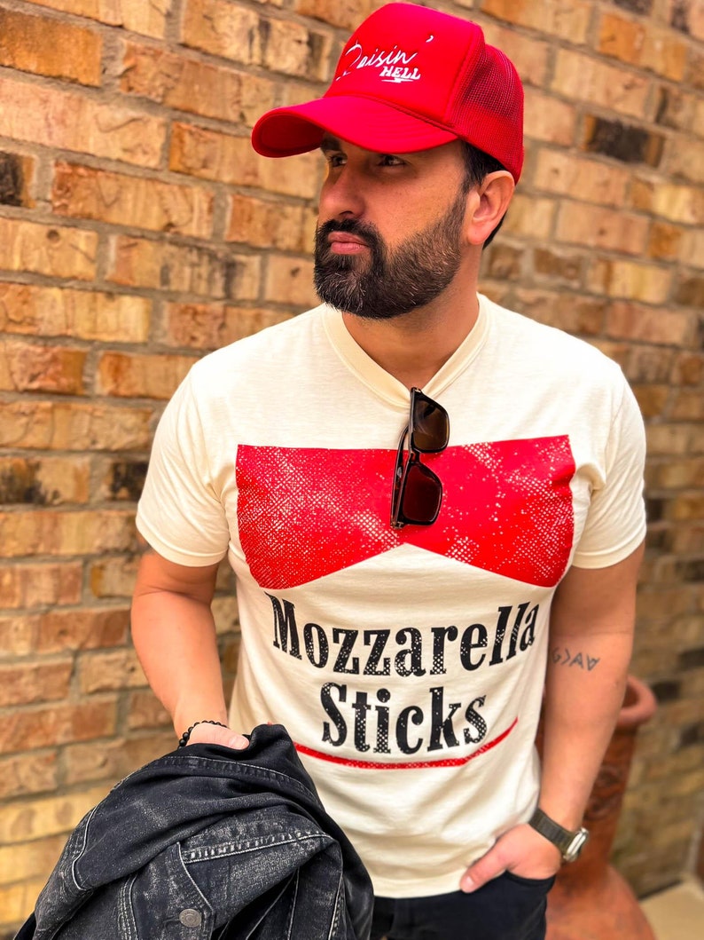 May include: A man wearing a red mesh trucker hat with the text "Cruisin' Hell" in white. He is wearing a white t-shirt with a red and white graphic that says "Mozzarella Sticks". He is also wearing a black leather jacket and black trousers.