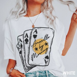 May include: White t-shirt with a graphic of four playing cards stacked on top of each other. The cards are black and white with a yellow circle in the center that says "Amarillo by Morning".