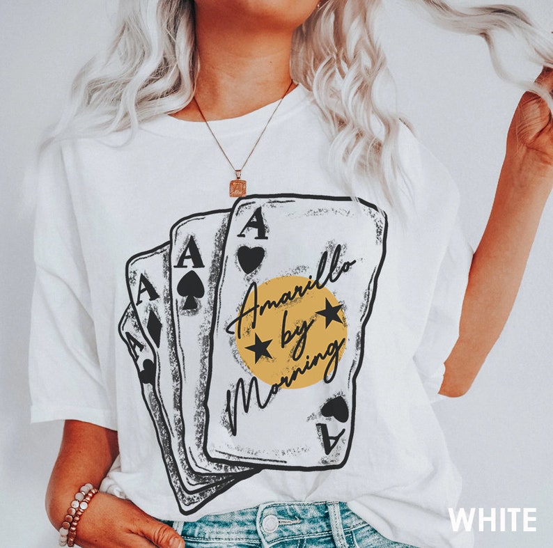 May include: White t-shirt with a graphic of four playing cards stacked on top of each other. The cards are black and white with a yellow circle in the center that says "Amarillo by Morning".