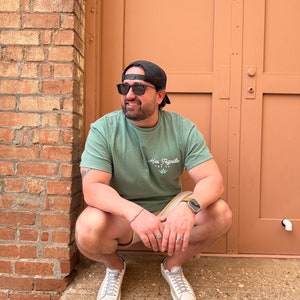 May include: A man wearing a green t-shirt with the text "Mas Tequila" and a graphic of a cactus, black sunglasses, a black baseball cap, and beige shorts. He is sitting on a brick surface with a brown door behind him.