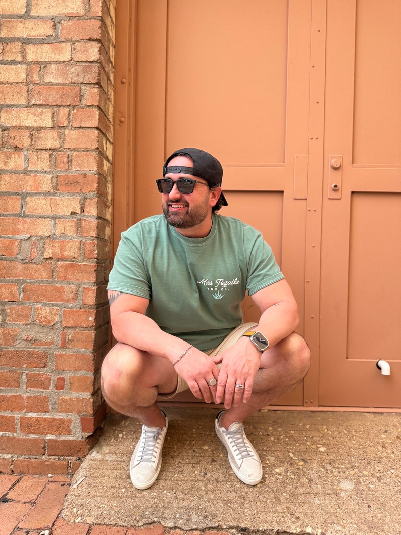 May include: A man wearing a green t-shirt with the text "Mas Tequila" and a graphic of a cactus, black sunglasses, a black baseball cap, and beige shorts. He is sitting on a brick surface with a brown door behind him.