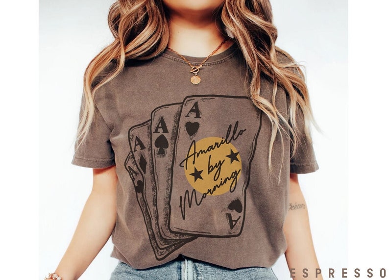 May include: A brown t-shirt with a graphic of four playing cards. The cards are arranged in a stack with the ace of spades on top. The graphic also includes a yellow circle with the text 'Amarillo by Morning' and two stars.