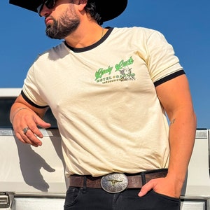 Puede incluir: Una persona lleva una camiseta de color crema con un ribete negro y un gr&aacute;fico verde que dice "Lady Luck Hotel + Casino" con una mujer con un sombrero de vaquero y una estrella. La persona lleva un cintur&oacute;n de cuero marr&oacute;n con una hebilla plateada y pantalones vaqueros negros. La persona lleva un sombrero de vaquero negro y gafas de sol.