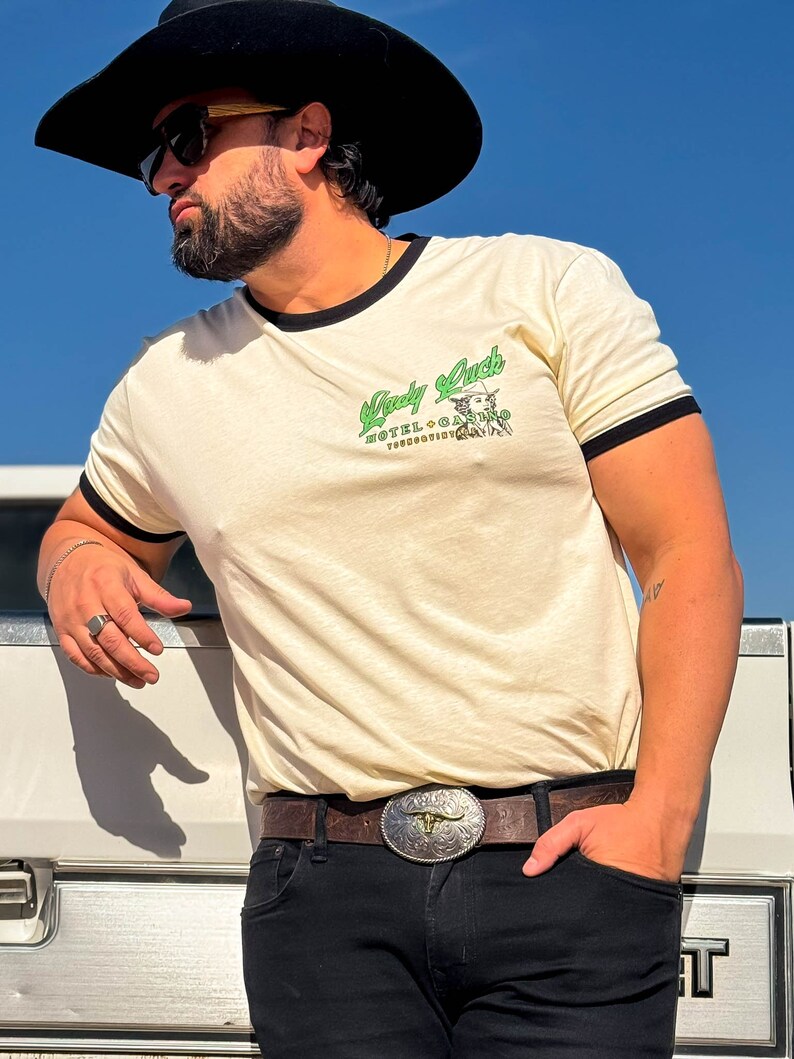 Puede incluir: Una persona lleva una camiseta de color crema con un ribete negro y un gr&aacute;fico verde que dice "Lady Luck Hotel + Casino" con una mujer con un sombrero de vaquero y una estrella. La persona lleva un cintur&oacute;n de cuero marr&oacute;n con una hebilla plateada y pantalones vaqueros negros. La persona lleva un sombrero de vaquero negro y gafas de sol.