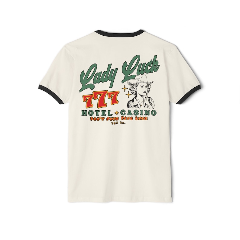 Puede incluir: Una camiseta blanca con un ribete negro. La parte posterior de la camiseta tiene un gr&aacute;fico de estilo vintage de una mujer con un sombrero de vaquero con el texto "Lady Luck 777 Hotel + Casino Don't Push Your Luck Too Far."