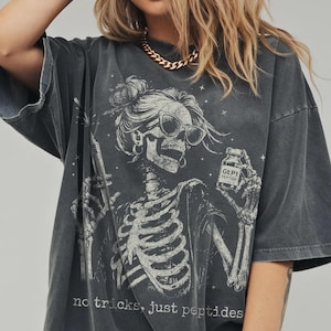 May include: A charcoal gray oversized t-shirt featuring a white graphic of a skeleton wearing sunglasses and a hair bun. The skeleton holds a syringe and a bottle labeled GLP1 PEPTIDE. The text below reads, no tricks, just peptides.