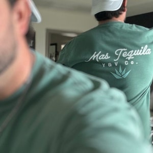 May include: Two people wearing green t-shirts with white hats. The person at the back has a white t-shirt with the text "Mas Tequila Y & V Co." printed on the back.