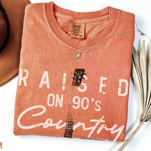 May include: A coral orange t-shirt with the text "RAISED ON 90'S Country" in white. A black and white guitar graphic is in the centre of the shirt.