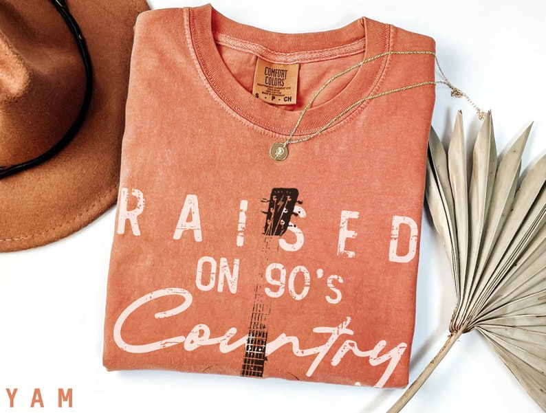 May include: A coral orange t-shirt with the text "RAISED ON 90'S Country" in white. A black and white guitar graphic is in the centre of the shirt.