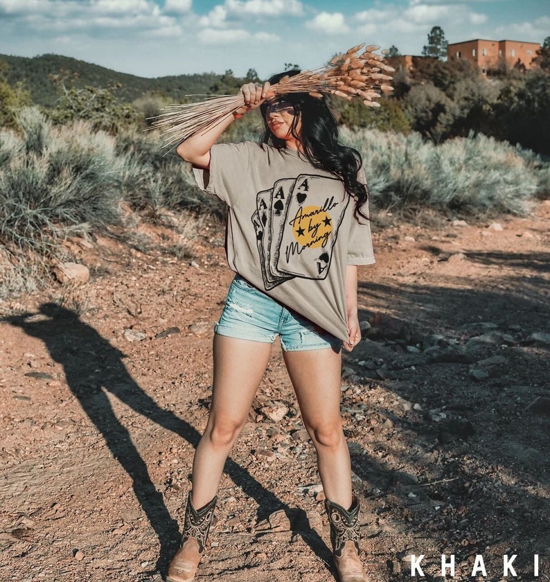 May include: A woman wearing a khaki t-shirt with a graphic of playing cards and the text "Amarillo by Morning". She is wearing denim shorts and cowboy boots. The photo was taken outdoors in a desert setting.