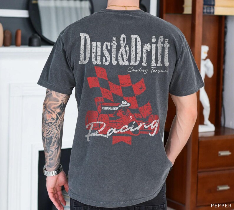 May include: Dark gray t-shirt with a distressed graphic design. The design features the words "Dust & Drift" above a red and black checkered flag and the word "Racing". The shirt has short sleeves.