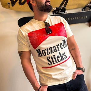 May include: A man wearing a red baseball cap with a white t-shirt that says "Mozzarella Sticks" in black and red lettering. The shirt has a red and white graphic design that resembles a box of mozzarella sticks.