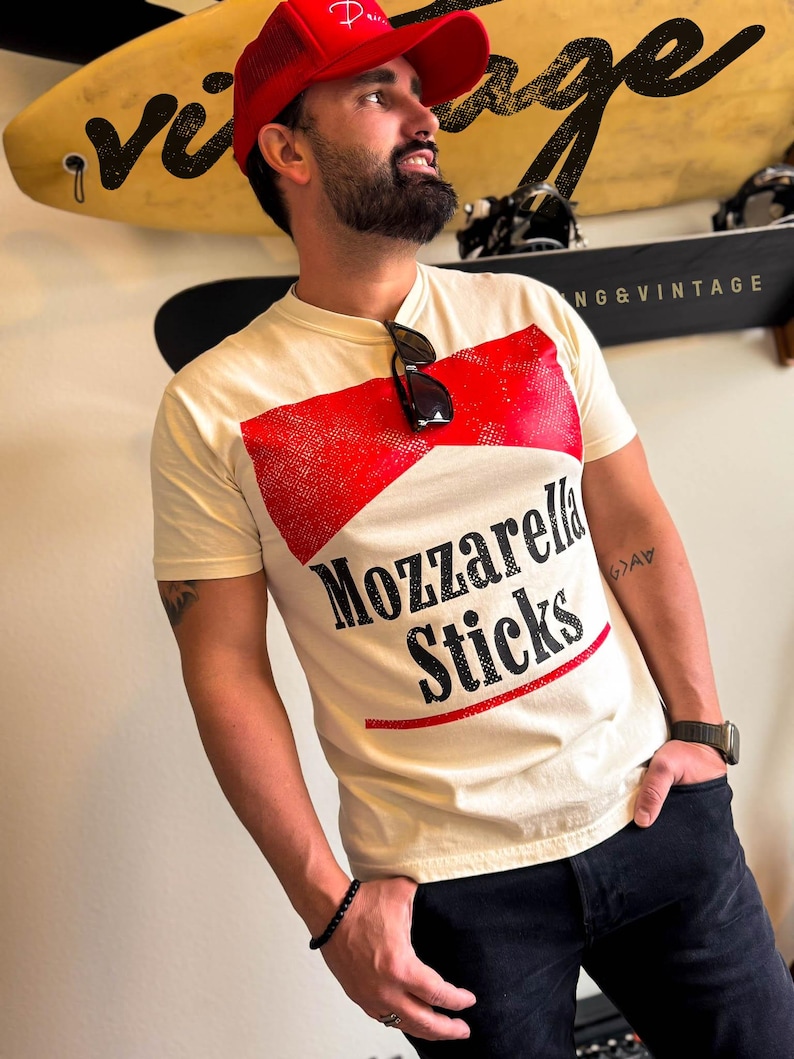 May include: A man wearing a red baseball cap with a white t-shirt that says "Mozzarella Sticks" in black and red lettering. The shirt has a red and white graphic design that resembles a box of mozzarella sticks.