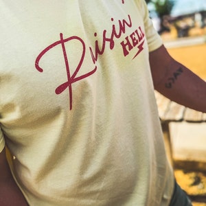 Peut inclure: Un t-shirt jaune clair avec un motif rouge qui dit "Risin' Hell" avec un &eacute;clair.