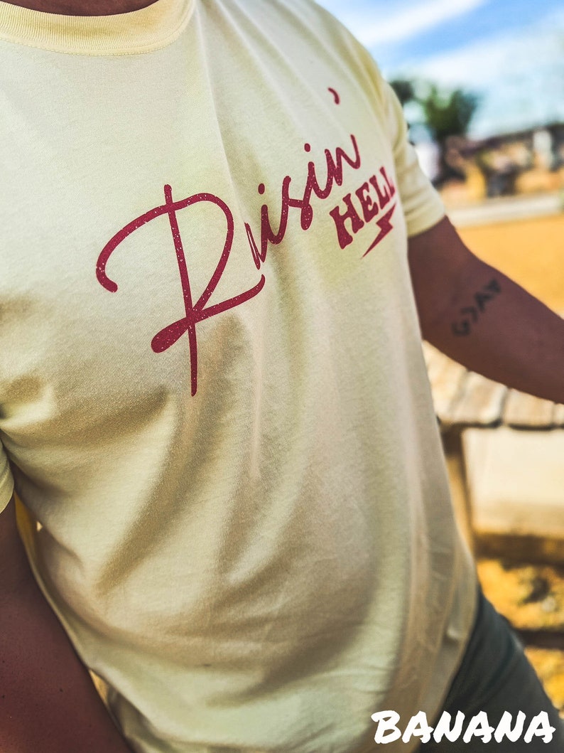 Peut inclure: Un t-shirt jaune clair avec un motif rouge qui dit "Risin' Hell" avec un &eacute;clair.