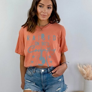 May include: A terracotta orange cropped t-shirt with a blue graphic that says "Raised on 90's Country" with a guitar and stars.