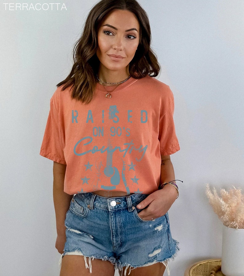 May include: A terracotta orange cropped t-shirt with a blue graphic that says "Raised on 90's Country" with a guitar and stars.