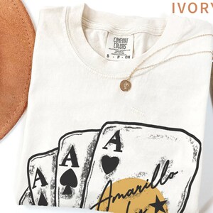 May include: Ivory t-shirt with a graphic of four playing cards, three of which are aces. The cards are arranged in a fan shape with the ace of hearts on top. The text "Amarillo" is written in a cursive font below the cards. A yellow sun with a star is behind the text.
