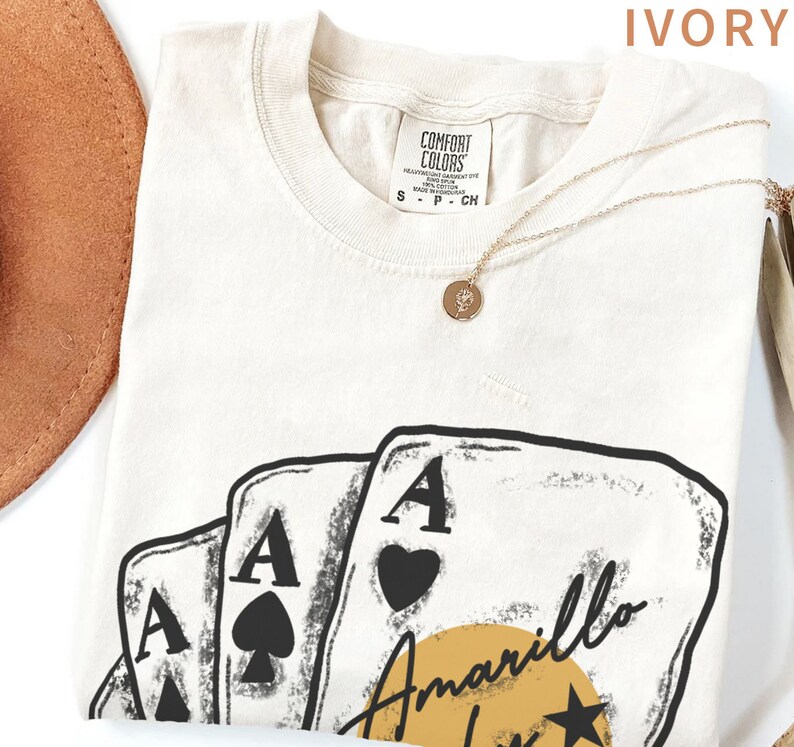 May include: Ivory t-shirt with a graphic of four playing cards, three of which are aces. The cards are arranged in a fan shape with the ace of hearts on top. The text "Amarillo" is written in a cursive font below the cards. A yellow sun with a star is behind the text.