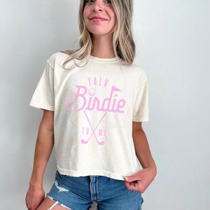 May include: A woman wearing a cream colored t-shirt with a pink graphic that says "Talk Birdie To Me" and a golf club design. She is wearing blue denim shorts.