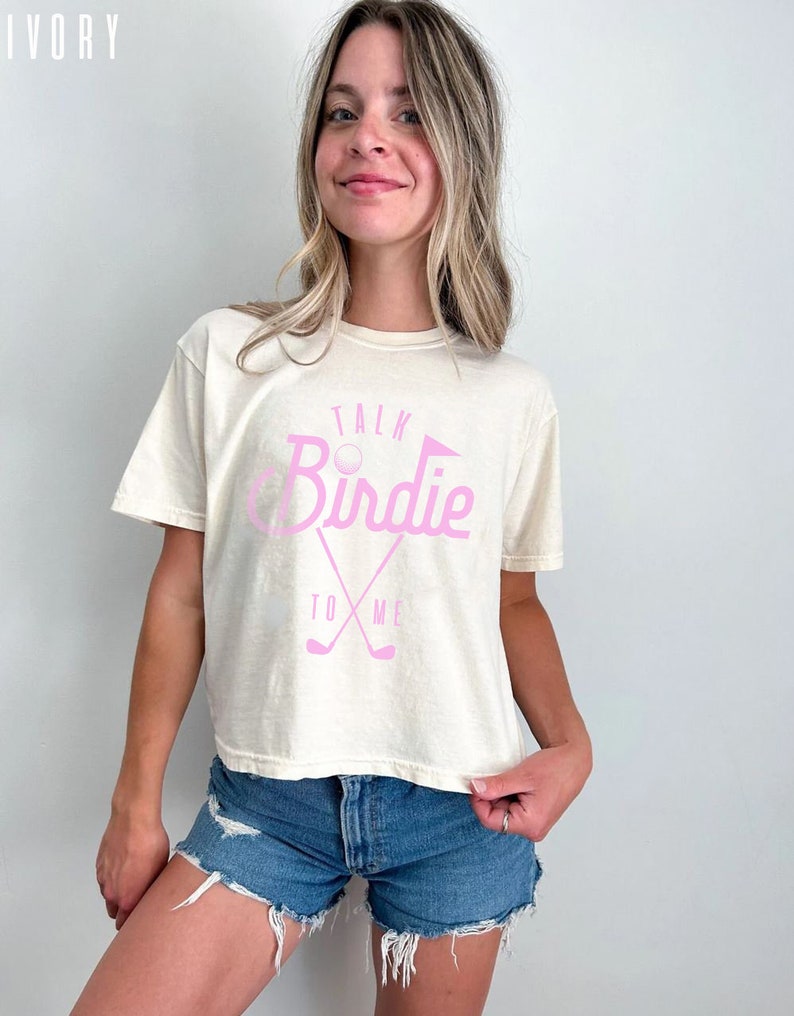 May include: A woman wearing a cream colored t-shirt with a pink graphic that says "Talk Birdie To Me" and a golf club design. She is wearing blue denim shorts.