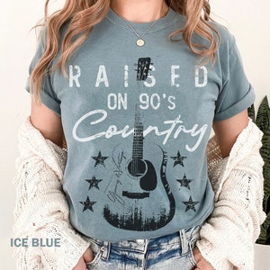 May include: A light blue t-shirt with the text "RAISED ON 90'S COUNTRY" in white. A black and white silhouette of a guitar is in the centre of the shirt with stars around it.