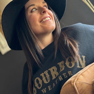 May include: A woman wearing a black sweatshirt with the text "Bourbon Weather" in gold lettering. She is wearing a black wide-brimmed hat and looking up.