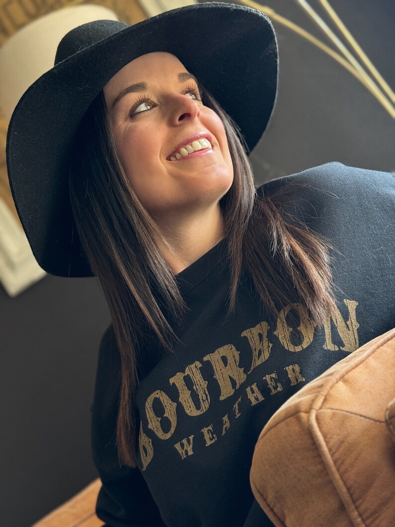 May include: A woman wearing a black sweatshirt with the text "Bourbon Weather" in gold lettering. She is wearing a black wide-brimmed hat and looking up.