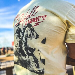 Peut inclure: Un t-shirt jaune clair avec une image en noir et blanc d'un cow-boy &agrave; cheval. Le texte "This Hell I Won't" est imprim&eacute; en rouge sur le t-shirt.