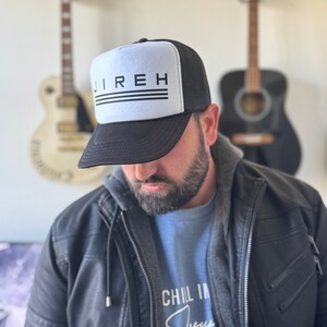 May include: A black and white trucker hat with the word "JIREH" printed in black on the front. The hat has a black mesh back and a white front panel. The person wearing the hat is also wearing a blue shirt with the words "CHIL IN Jesus" printed on it.