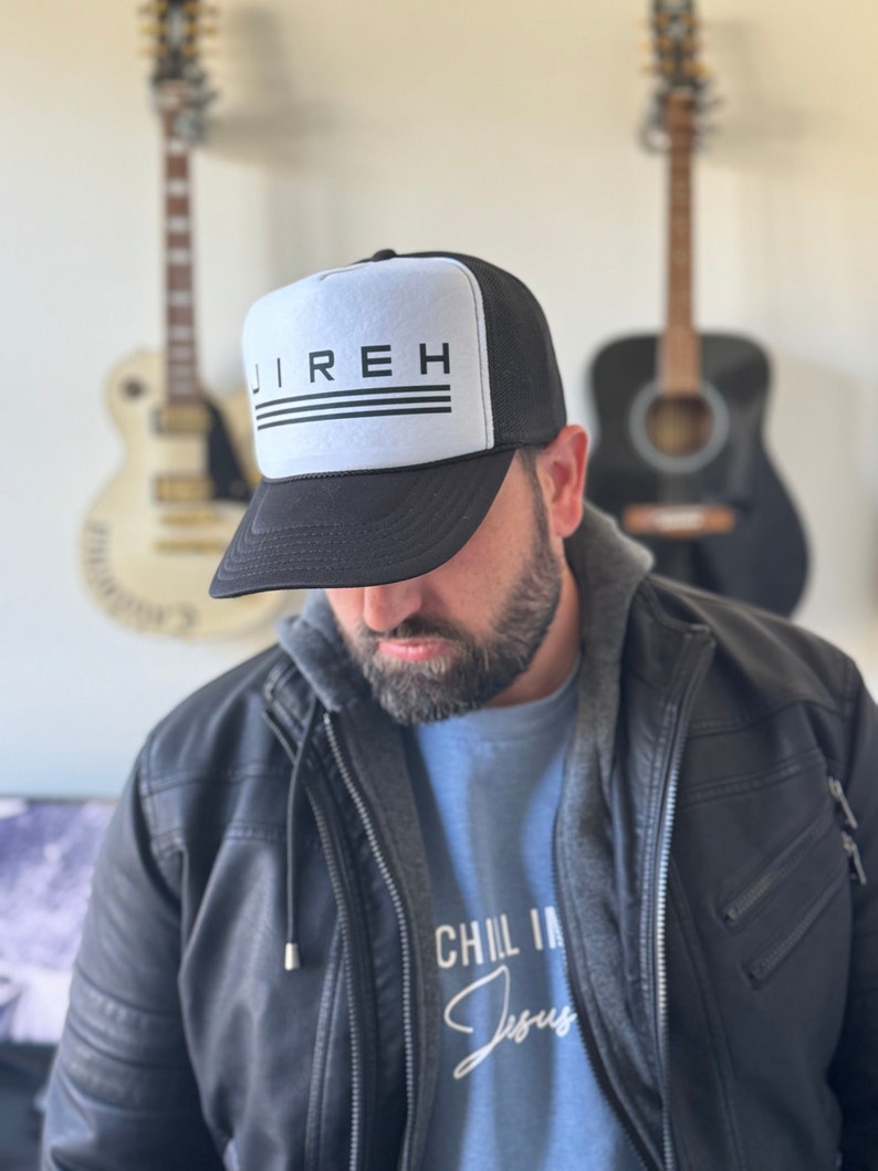 May include: A black and white trucker hat with the word "JIREH" printed in black on the front. The hat has a black mesh back and a white front panel. The person wearing the hat is also wearing a blue shirt with the words "CHIL IN Jesus" printed on it.