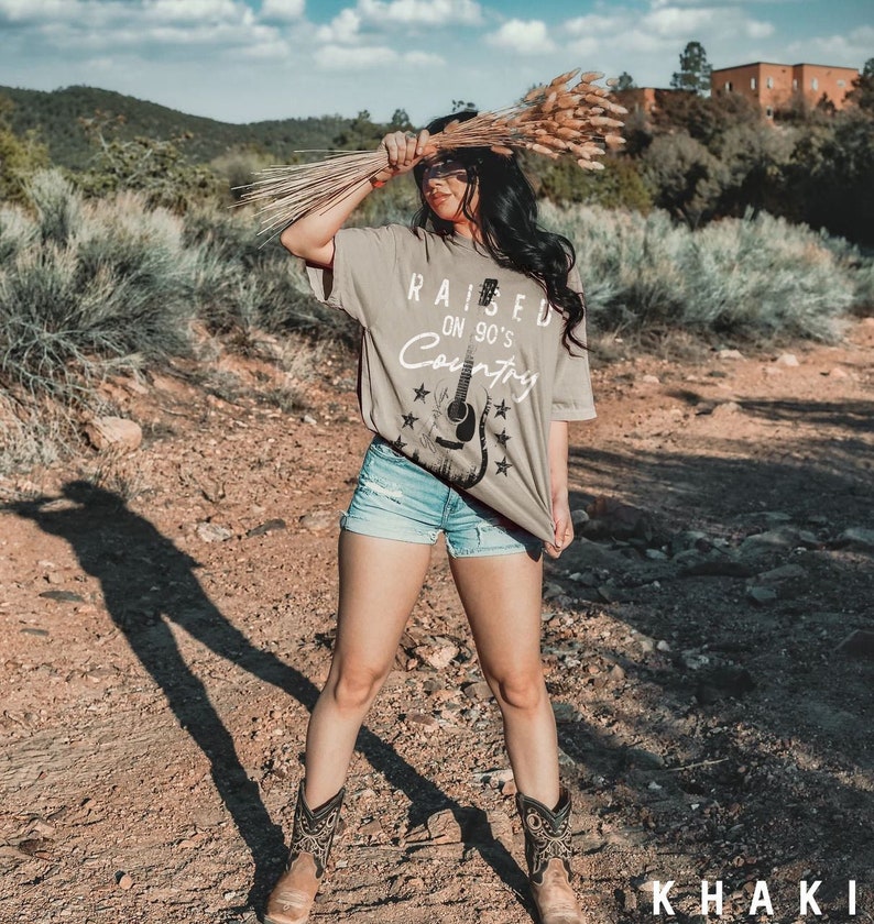 May include: A woman wearing a khaki t-shirt with the text "Raised on 90's Country" and a guitar graphic. She is wearing denim shorts and brown cowboy boots. The photo was taken outdoors in a desert setting.