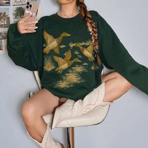May include: Dark green sweatshirt featuring a golden illustration of ducks in flight over water. The long-sleeved top has a crew neck and a relaxed fit. The ducks are detailed with brown and tan accents, creating a nature-inspired design.