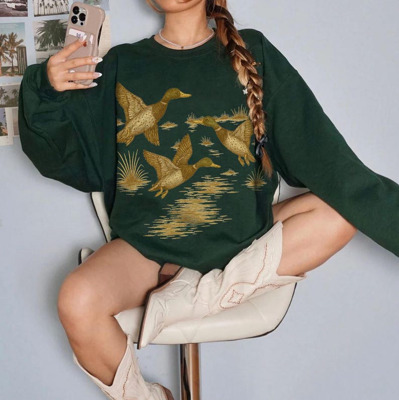 May include: Dark green sweatshirt featuring a golden illustration of ducks in flight over water. The long-sleeved top has a crew neck and a relaxed fit. The ducks are detailed with brown and tan accents, creating a nature-inspired design.