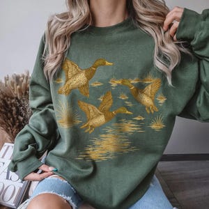May include: Green sweatshirt featuring a detailed illustration of three ducks in flight over a body of water. The ducks are depicted in shades of brown and gold, with the water and reeds also in gold. The sweatshirt is a solid green color.